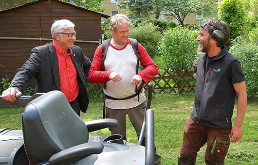 Drei Männer unterhalten sich lachend im Garten neben einem Aufsitzrasenmäher; einer trägt Arbeitskleidung und Gehörschutz, einer einen Laubbläser, der ältere im Sakko lacht.